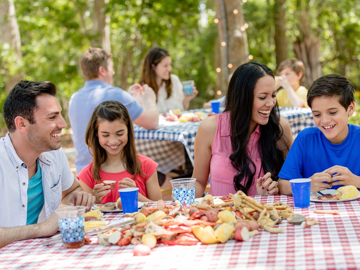Families laughing and eating at picnic tables outdoors sharing family vacation moments. 
